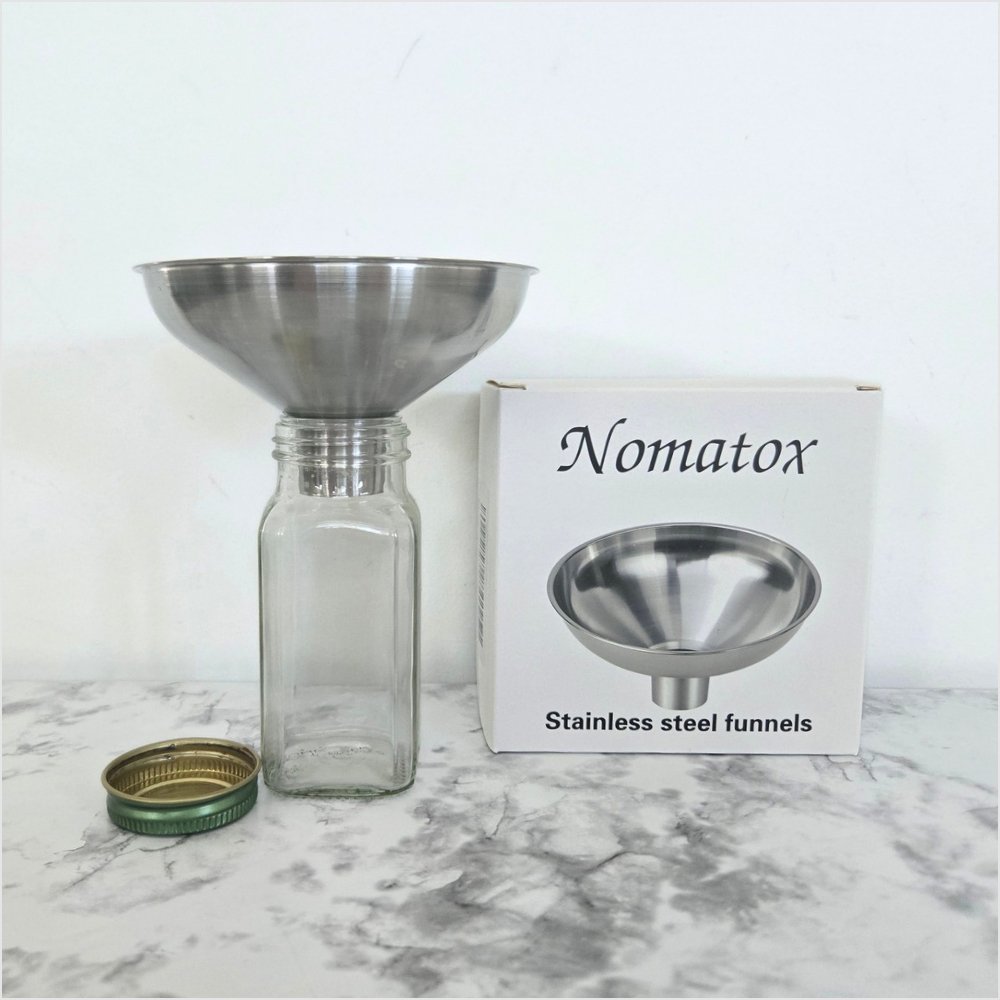 Nomatox stainless steel funnel resting in an empty glass spice jar beside its box and the jar’s lid on a marble counter, for mess-free refills of bulk organic spices.